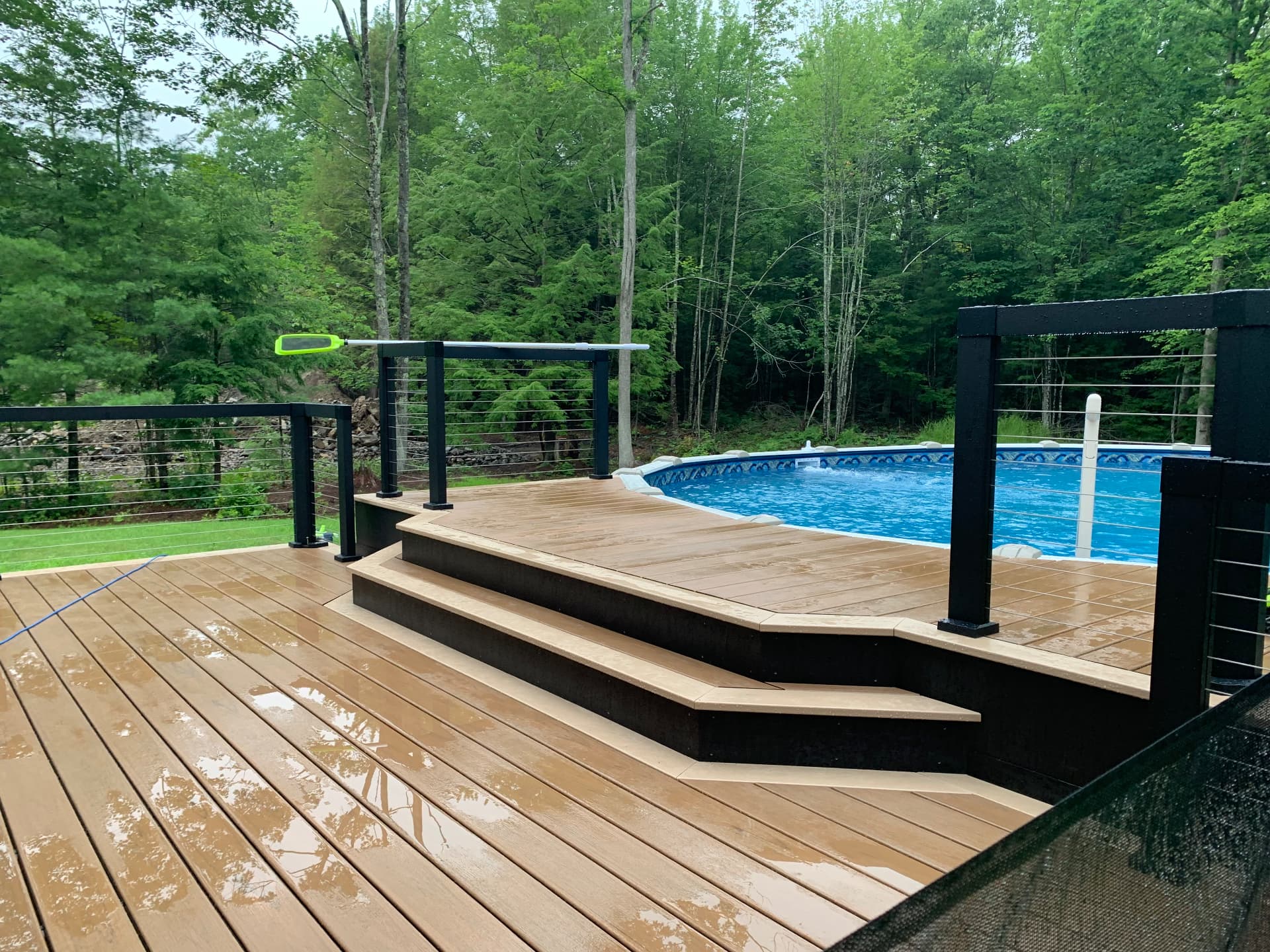 Multi-level brown composite pool deck with black cable railings around an above-ground pool